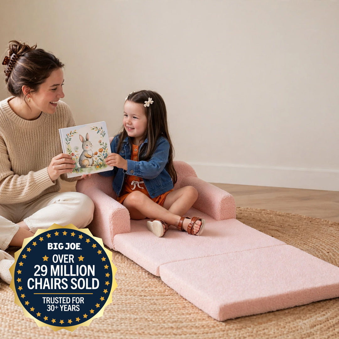 A woman and a young girl sit on a pink foldable floor chair in a bright room, reading a book together. The chair is upholstered in a soft blush pink fabric and has a built-in storage compartment. 