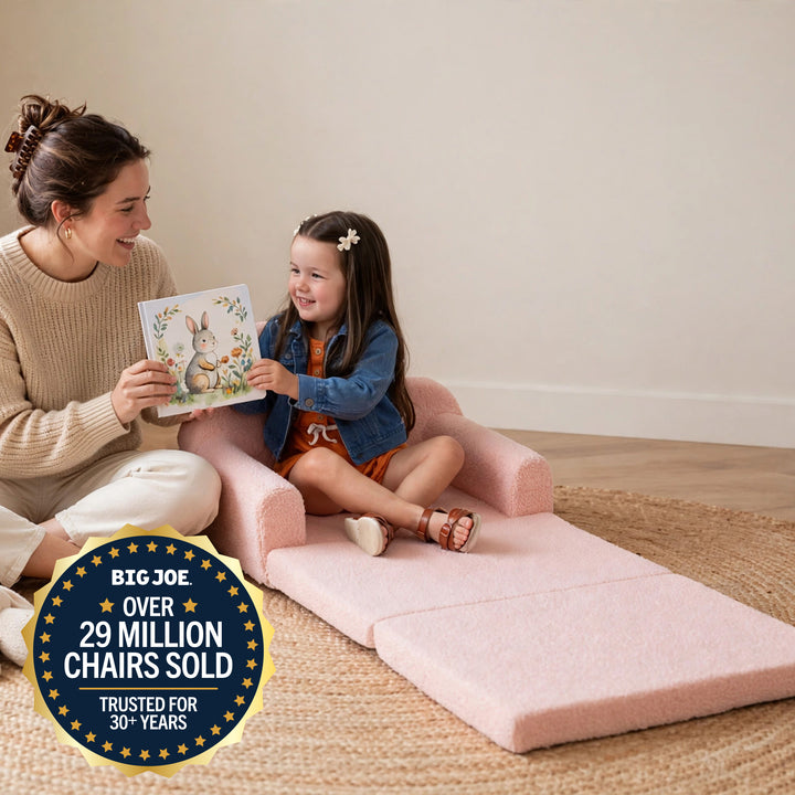 A woman and a young girl sit on a pink foldable floor chair in a bright room, reading a book together. The chair is upholstered in a soft blush pink fabric and has a built-in storage compartment. 