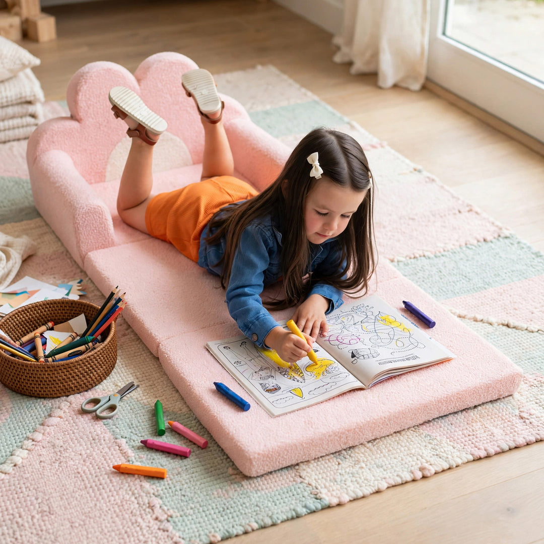 A young girl lies on a pink cloud-shaped foam play mat on a woven rug, coloring in a book with crayons scattered around in a bright room. 