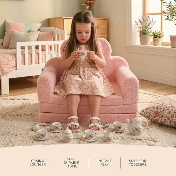 A young girl sits in a small pink boucle chair lounge in a cozy bedroom setting, holding a teacup from a matching floral tea set arranged on the floor. The scene includes text overlays at the bottom describing the chair and lounge features. 