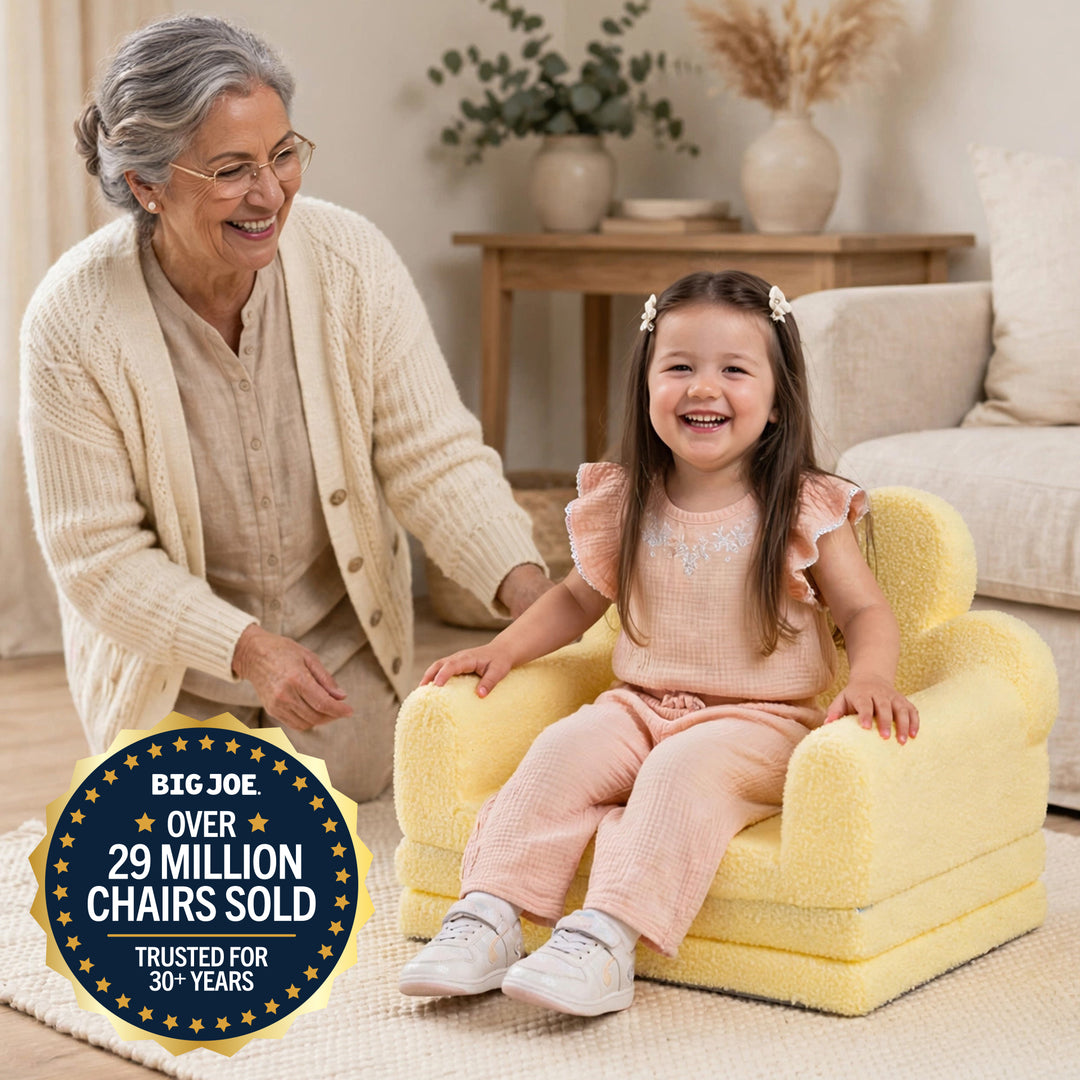 A young girl sits smiling in a small yellow plush armchair in a cozy living room, with an older woman kneeling beside her. A dark blue circular badge in the lower left reads &