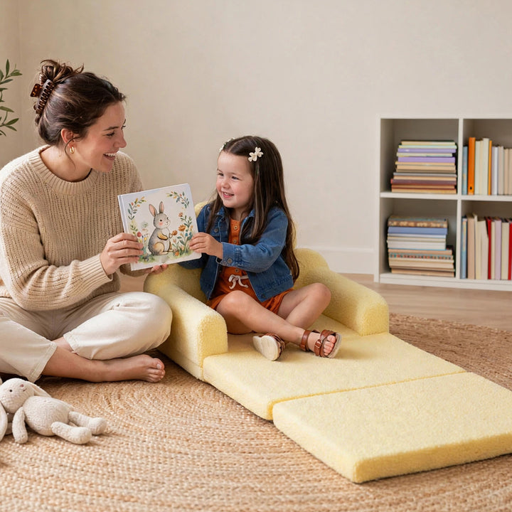 A woman and a young girl sit on a pale yellow foam floor cushion in a cozy room. The woman holds a children&