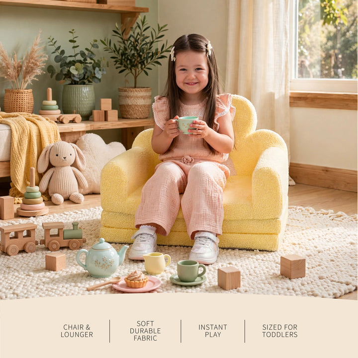 A young child sits in a small yellow plush toddler lounge chair in a styled nursery or playroom, holding a cup. The scene includes toys, plants, shelves, and a rug, with text at the bottom describing the chair features. 