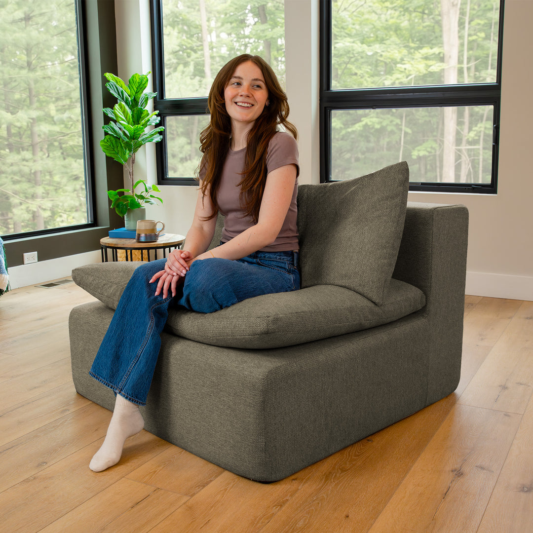 Woman sitting on a gray armchair in a room with large windows and a plant. 