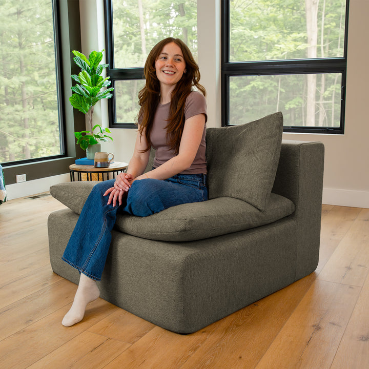 Woman sitting on a gray armchair in a room with large windows and a plant. 