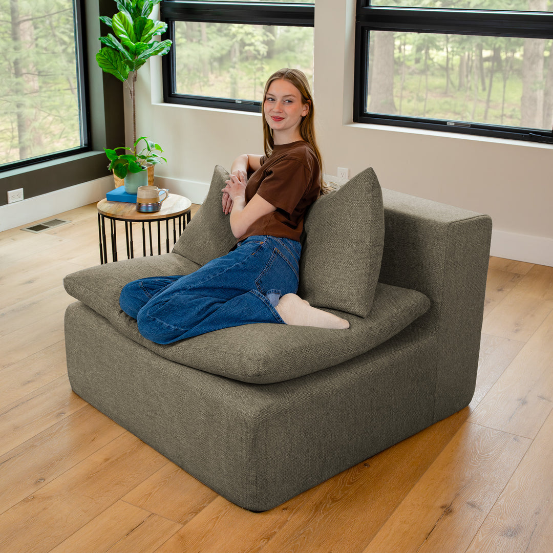 Person sitting on a gray sectional sofa in a room with large windows and plants. 