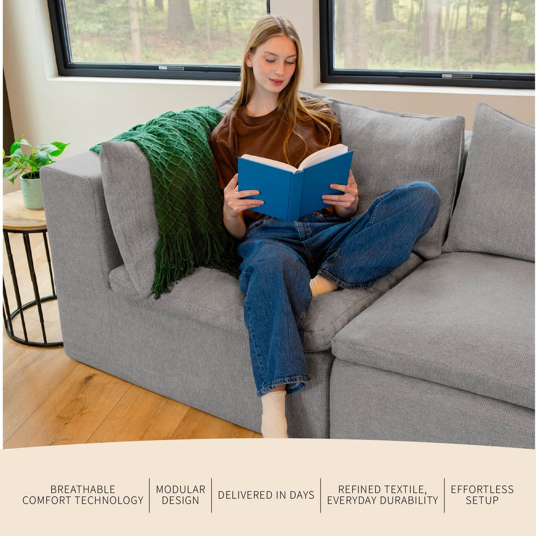 Woman reading a book on a gray couch in a bright room with large windows. 