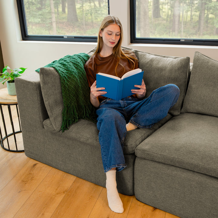 Woman reading a blue book on a gray couch in a room with large windows and plants. 