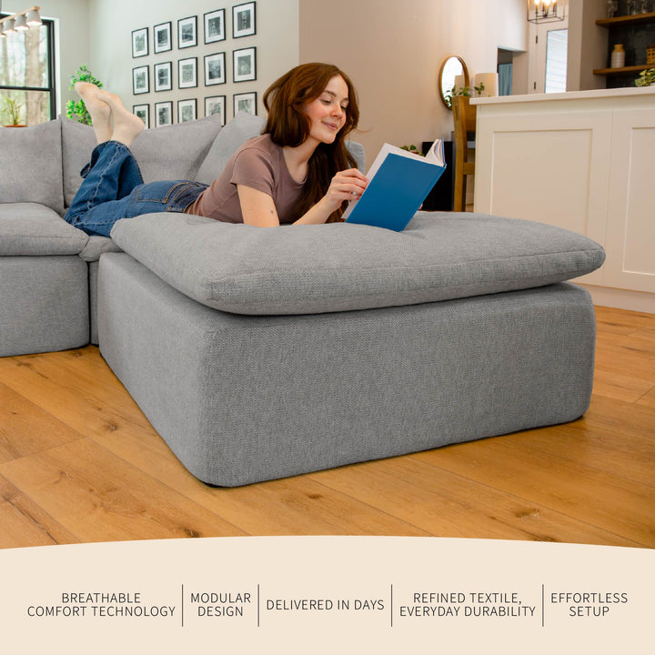 Woman reading a book on a large gray ottoman in a living room. 