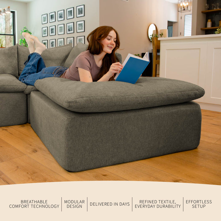 Woman reading a book on a large gray ottoman in a living room. 