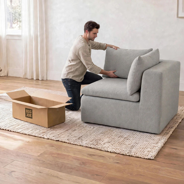 A man kneels beside a light gray upholstered armchair in a bright room, adjusting the back cushion. A cardboard box printed with &