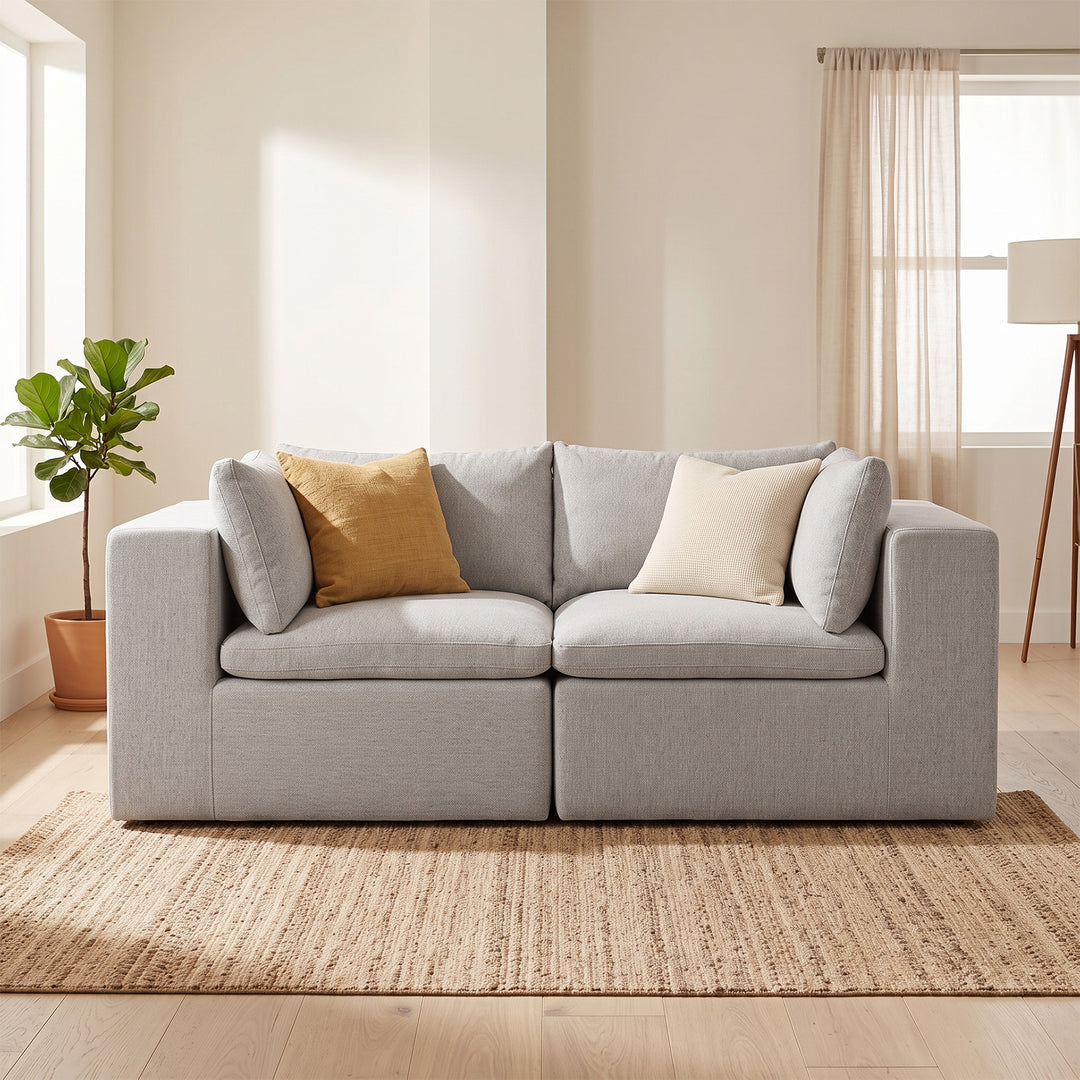 A light gray two-seater sofa with two accent pillows, one mustard yellow and one cream, set on a woven jute rug in a bright, modern living room. #color_cottage-gray-terra-knit
