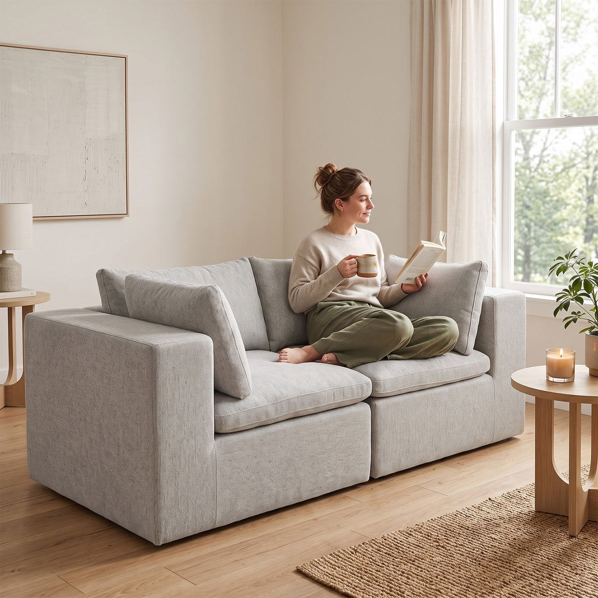 A woman sits barefoot on a light gray upholstered loveseat in a bright, modern living room, holding a book and mug. The room has light wood floors, a window with sheer curtains, and small wooden side tables. #color_cottage-gray-terra-knit