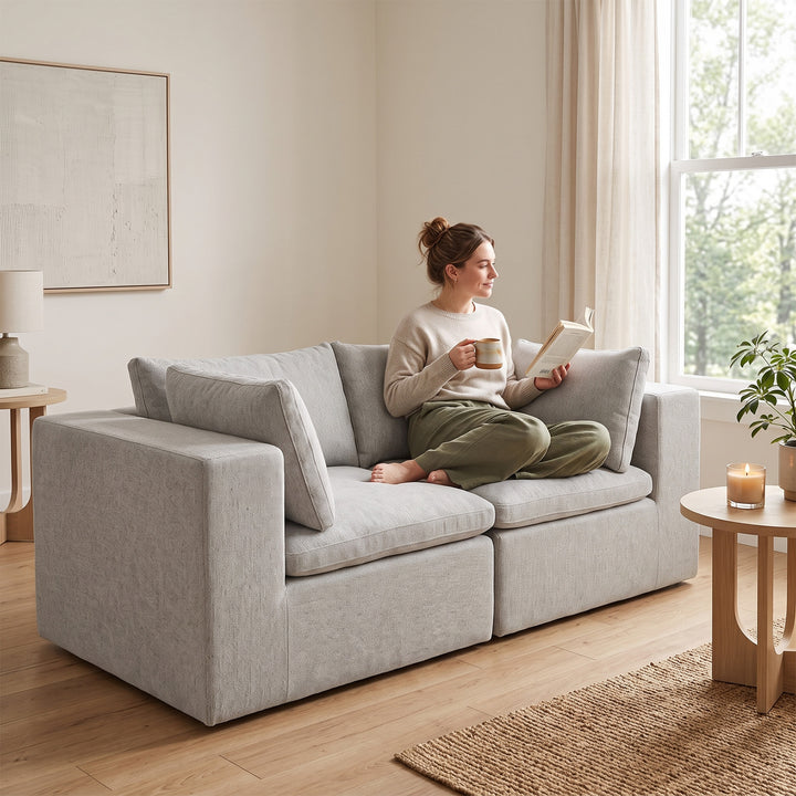A woman sits barefoot on a light gray upholstered loveseat in a bright, modern living room, holding a book and mug. The room has light wood floors, a window with sheer curtains, and small wooden side tables. 
