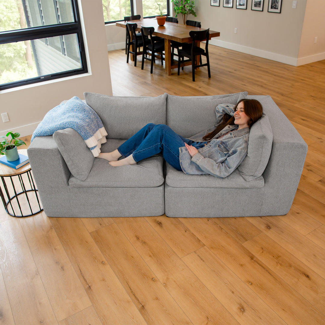 Woman sitting on a gray sectional sofa in a living room with a dining area in the background. 