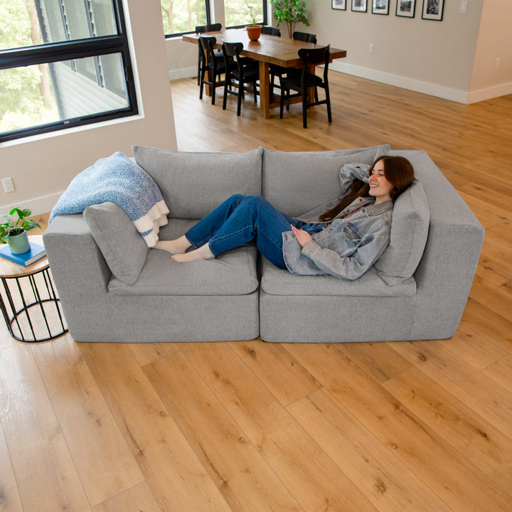Woman sitting on a gray sectional sofa in a living room with a dining area in the background. 
