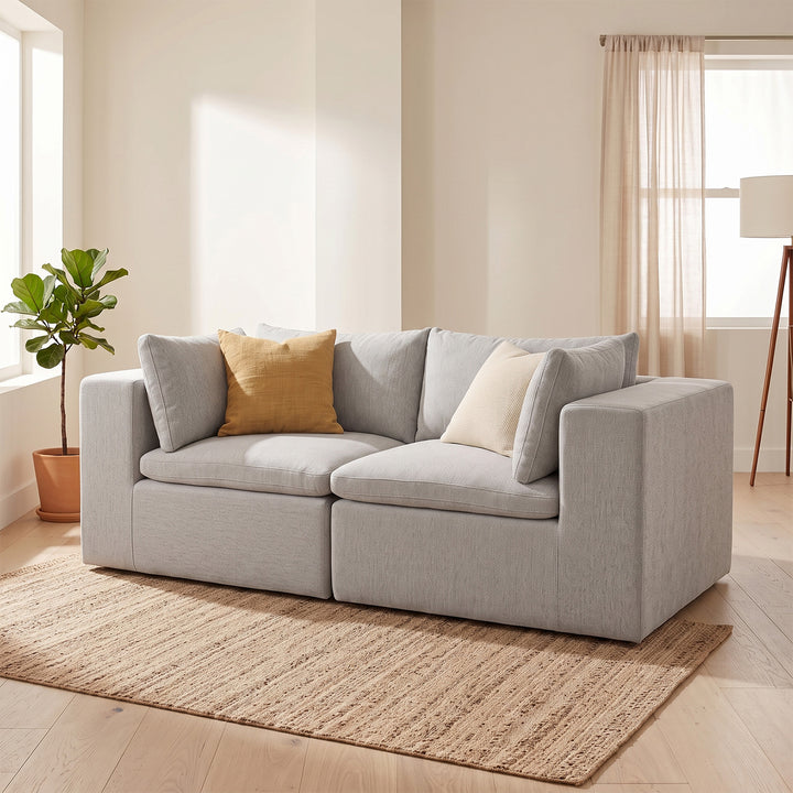 A light gray upholstered sofa with three throw pillows, including one mustard yellow pillow and one cream pillow, set on a woven jute rug in a bright, modern living room. 