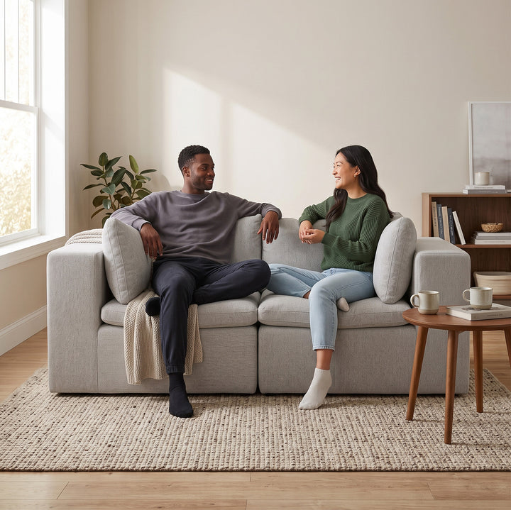 A man and a woman sit on a light gray modular sofa in a bright, modern living room, smiling and talking. The room has beige walls, a woven rug, a small wooden side table with mugs, and a bookshelf in the background. 