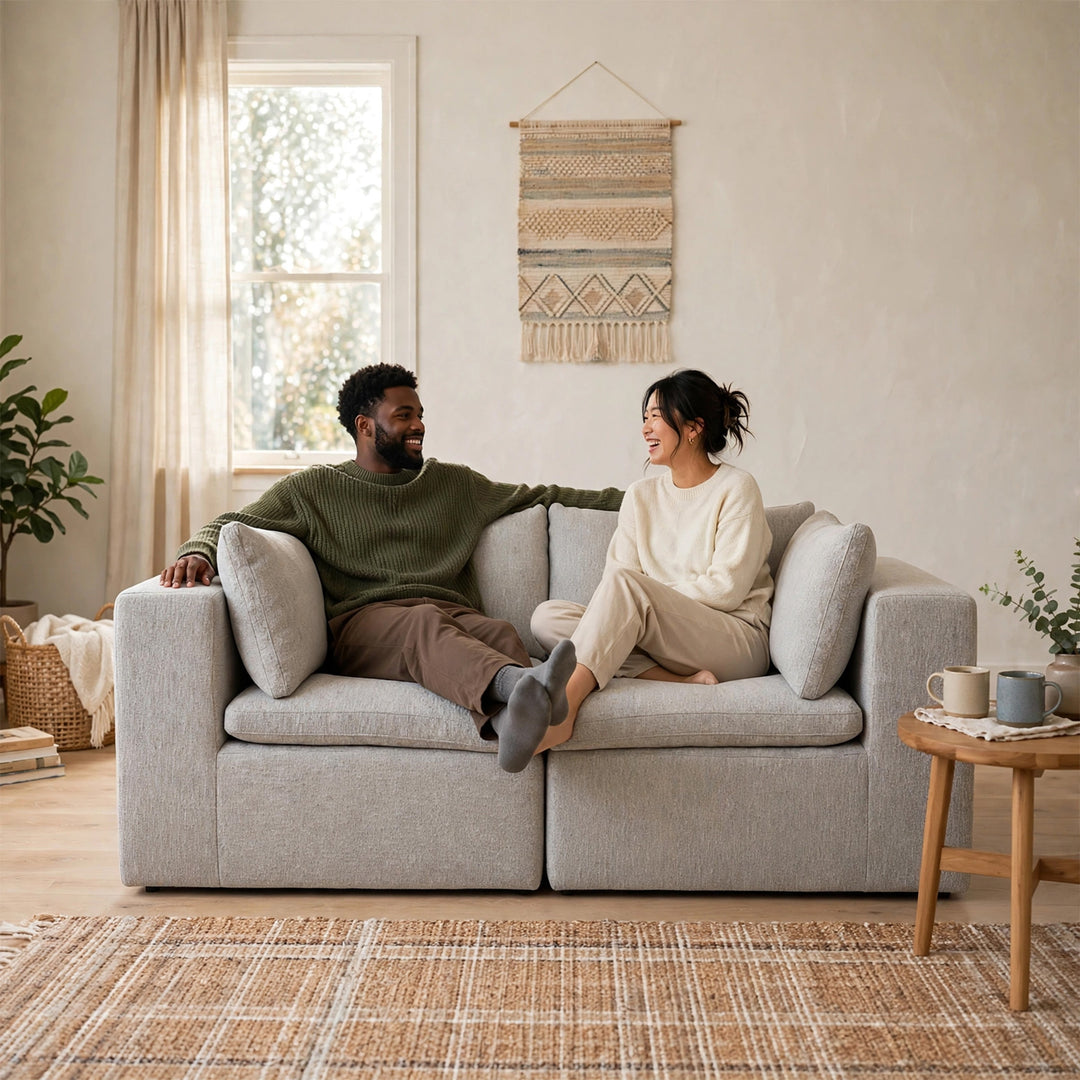 A man and a woman relax on a light gray modular two-seater sofa in a bright, modern living room with beige walls, a window, and a woven wall hanging. 