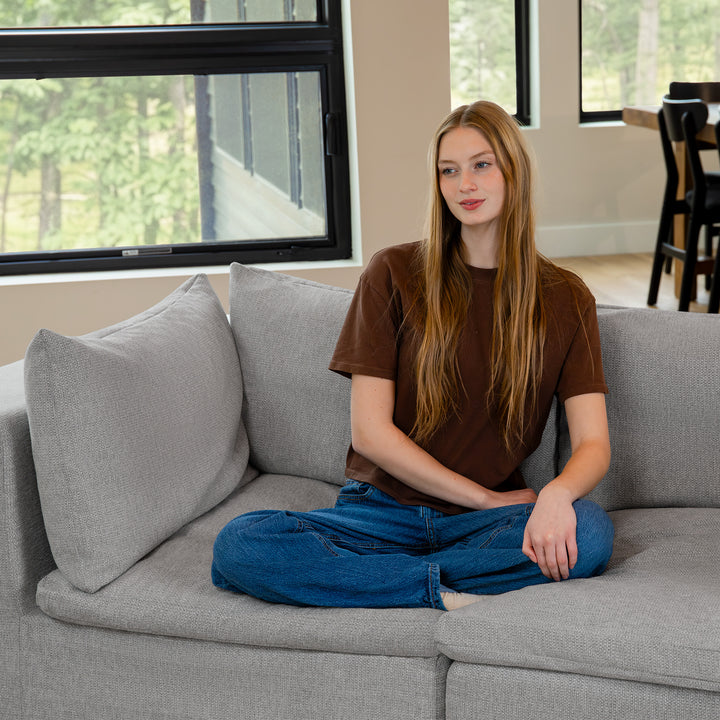 Person sitting on a gray couch in a room with large windows. 