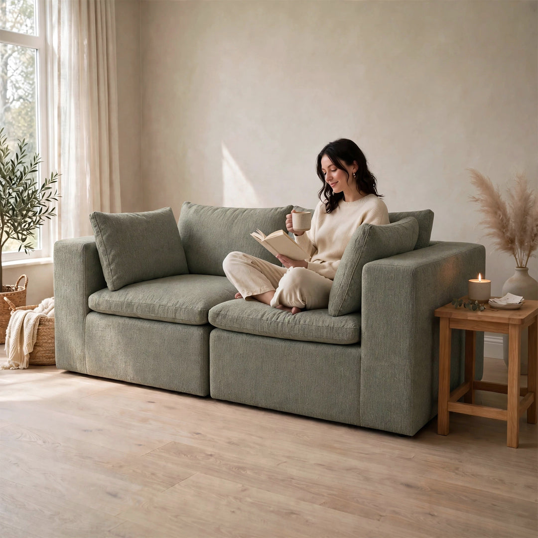 A woman sits on a large upholstered loveseat in a softly lit room, reading a book and holding a mug. The loveseat is a muted sage green with chunky armrests and matching back cushions. 