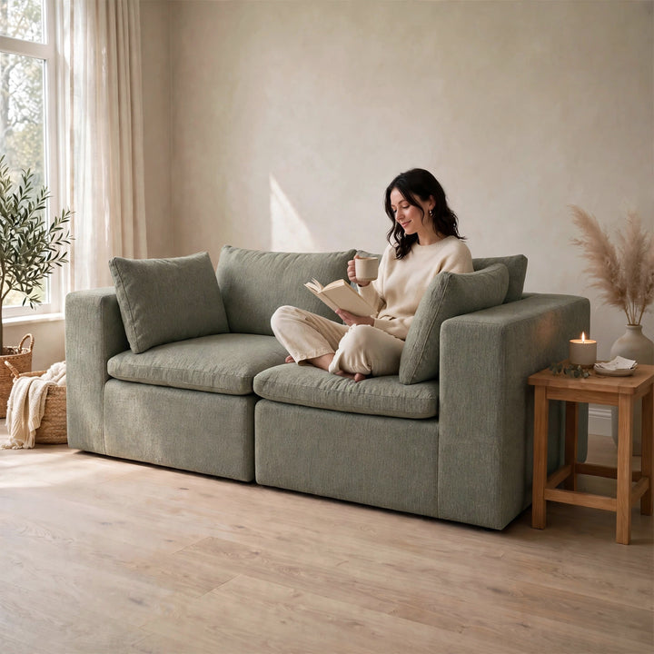 A woman sits on a large upholstered loveseat in a softly lit room, reading a book and holding a mug. The loveseat is a muted sage green with chunky armrests and matching back cushions. 