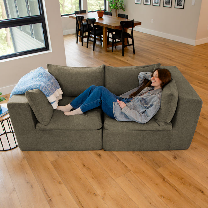 Woman sitting on a gray couch in a living room with a dining area in the background. 