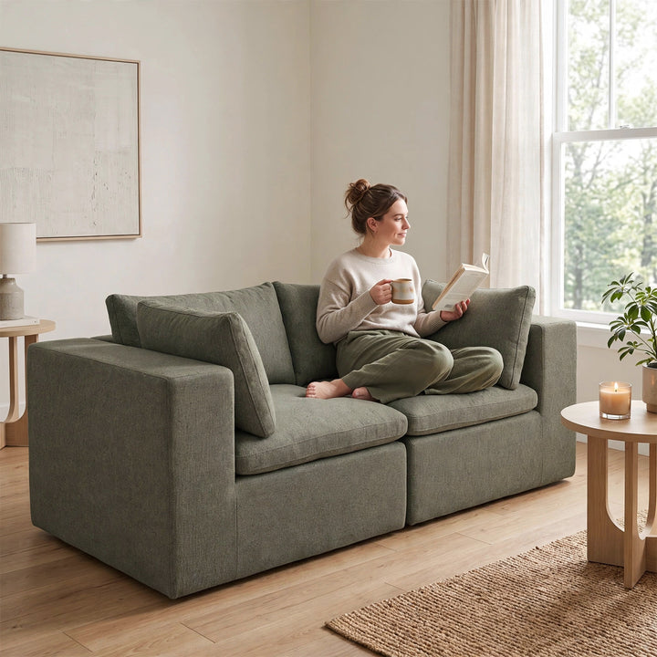 A woman sits cross-legged on a large, modern two-seat sofa in a bright, neutral-toned living room, holding a book and a mug. The sofa is upholstered in a textured sage green fabric. 