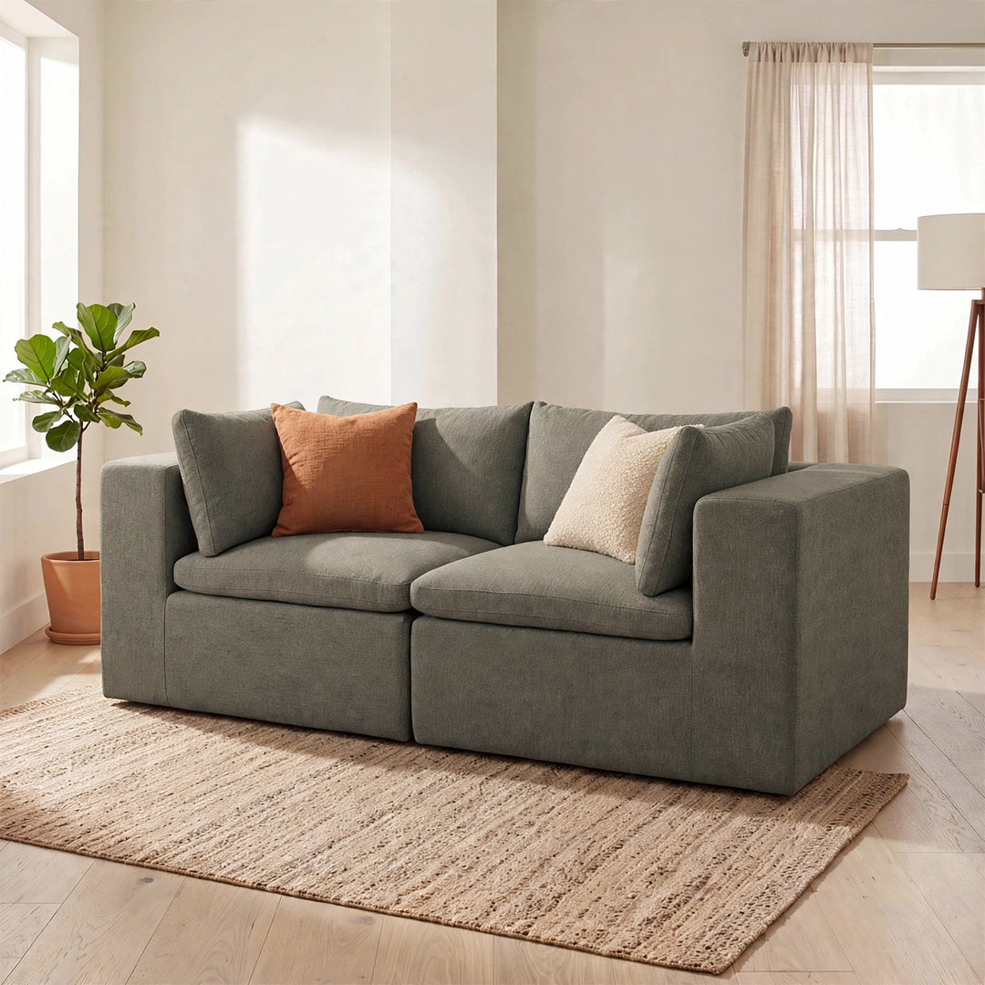 A modern two-seater sofa in textured gray fabric, styled with two accent pillows in a bright, minimalist living room with a woven rug, potted plant, and floor lamp. 