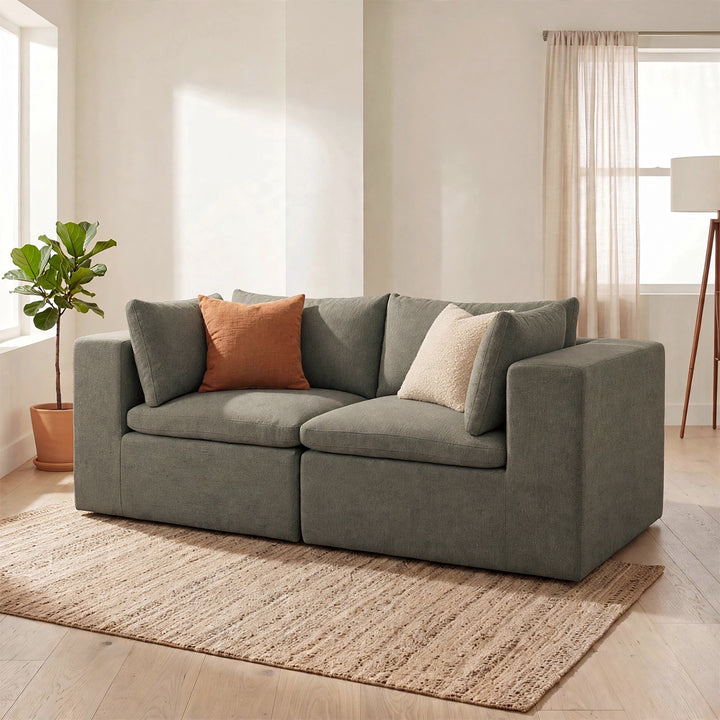 A modern two-seater sofa in textured gray fabric, styled with two accent pillows in a bright, minimalist living room with a woven rug, potted plant, and floor lamp. 