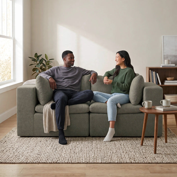 A man and a woman relax on a muted sage green two-seater sofa in a bright, modern living room. The man wears a gray long-sleeve shirt and dark pants, while the woman wears a green knit sweater and light blue jeans. 