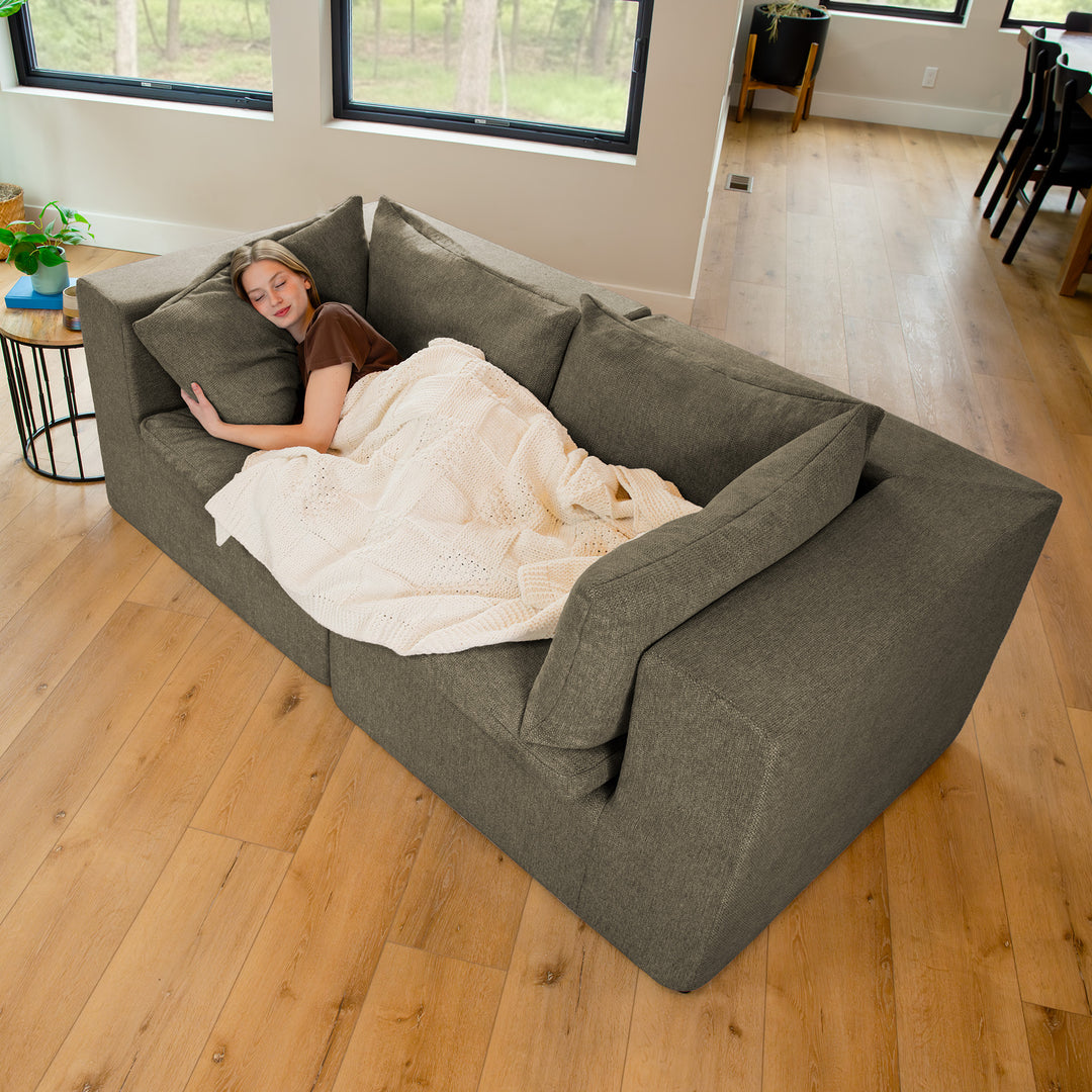 Woman lying on a large gray sofa in a room with wooden flooring and large windows. 