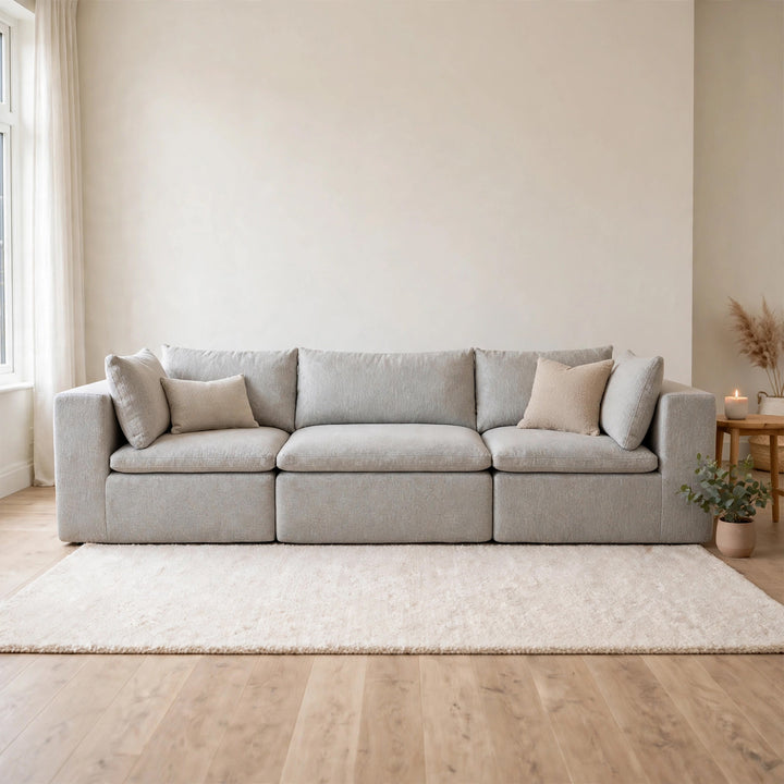 A light gray upholstered three-seat sofa with matching throw pillows, set in a bright, modern living room on a cream shag rug over light wood flooring. 
