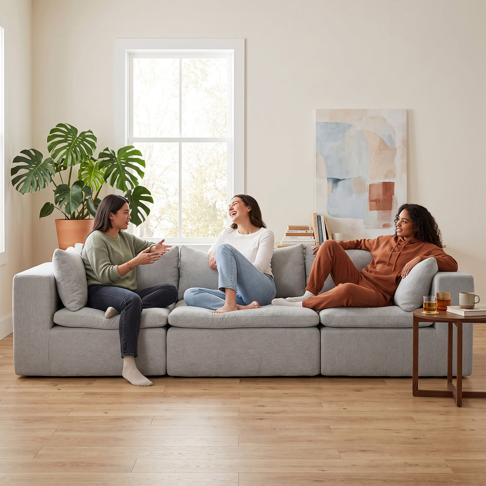 Three women sitting and chatting on a light gray modular sectional sofa in a bright, modern living room with wood floors, a potted plant, and a small side table. #color_cottage-gray-terra-knit