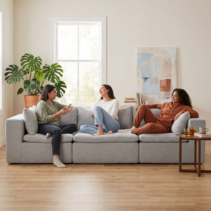 Three women sitting and chatting on a light gray modular sectional sofa in a bright, modern living room with wood floors, a potted plant, and a small side table. 