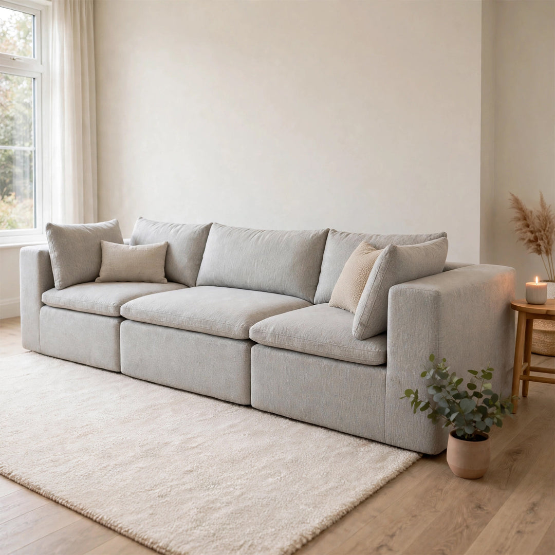 A light gray upholstered sofa with multiple seat and back cushions, styled in a bright living room with cream walls, a white rug, wood flooring, a small side table with a candle, and a potted plant. 