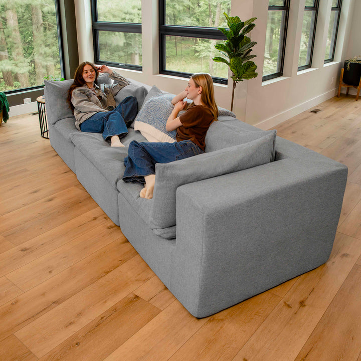 Two people sitting on a large gray sectional sofa in a room with large windows and wooden flooring. 