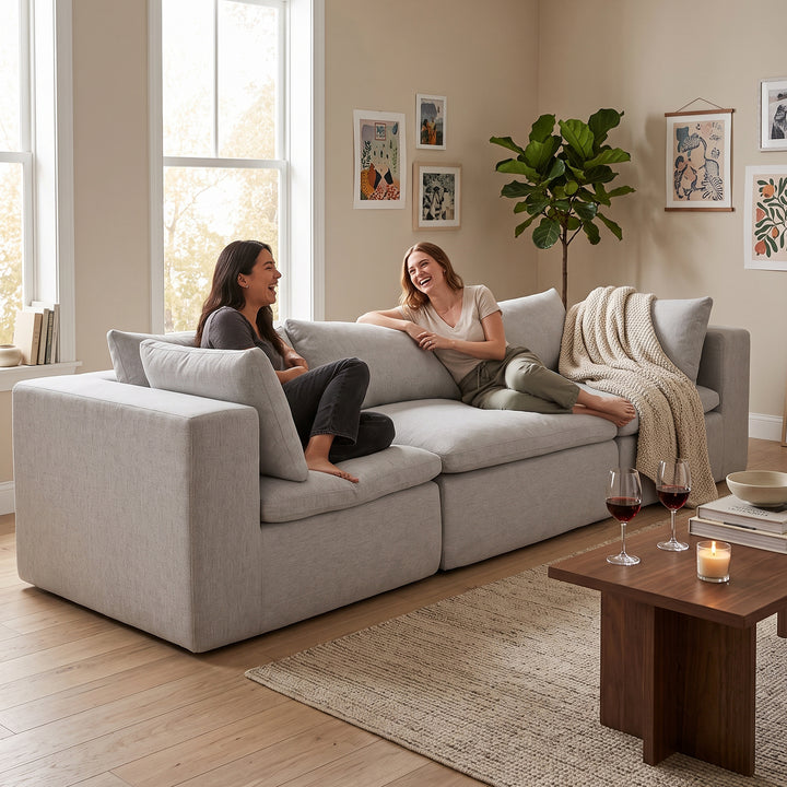 A light gray upholstered sectional sofa in a bright, modern living room with two women sitting and chatting on the couch. The room features beige walls, framed art, a potted plant, a wooden coffee table with wine glasses and a candle, and a woven rug. 