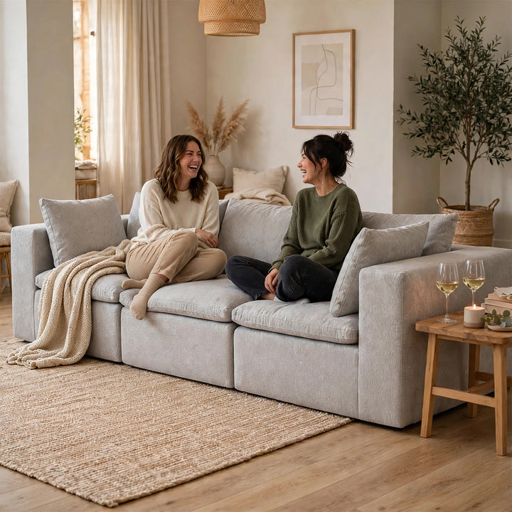 Two women laughing on a light gray modular sofa in a warm, modern living room with wood floors, a woven rug, and a small wooden side table with wine glasses. 