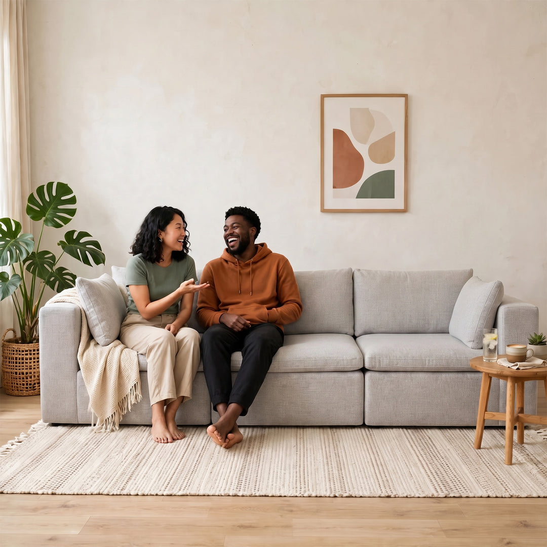 A man and a woman are sitting on a light gray fabric sofa in a bright, modern living room. The man wears an orange-brown hoodie and black pants, and the woman wears a sage green top and beige pants. 
