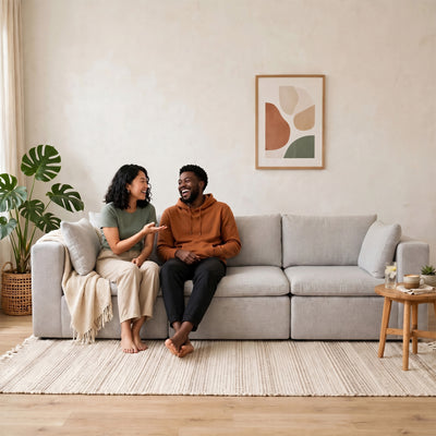 A man and a woman are sitting on a light gray fabric sofa in a bright, modern living room. The man wears an orange-brown hoodie and black pants, and the woman wears a sage green top and beige pants. #color_cottage-gray-terra-knit