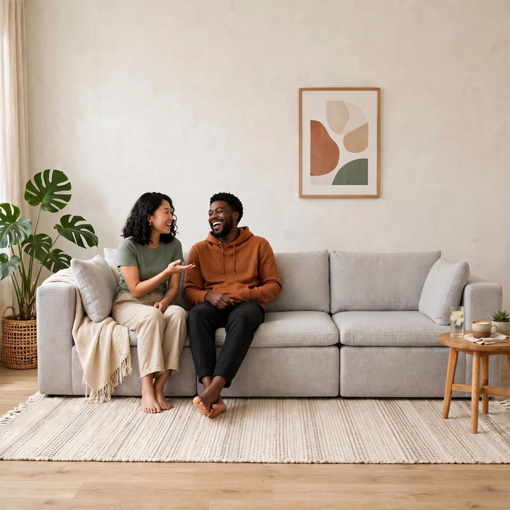 A man and a woman are sitting on a light gray fabric sofa in a bright, modern living room. The man wears an orange-brown hoodie and black pants, and the woman wears a sage green top and beige pants. 