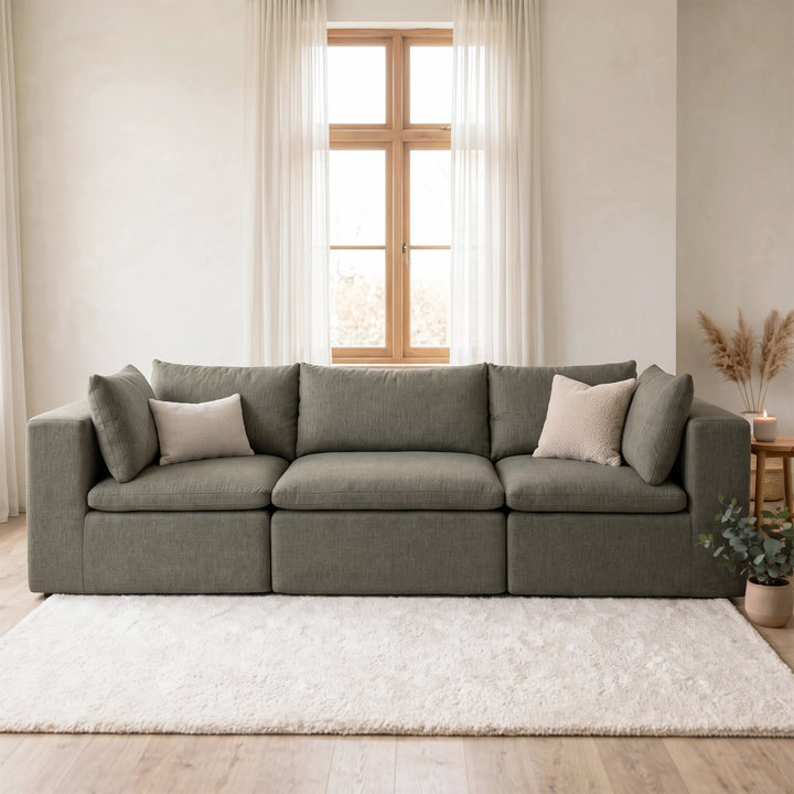 A modern three-seater sofa upholstered in textured gray fabric, styled with two light beige accent pillows, set in a bright, minimalist living room with a white shag rug and a window in the background. 