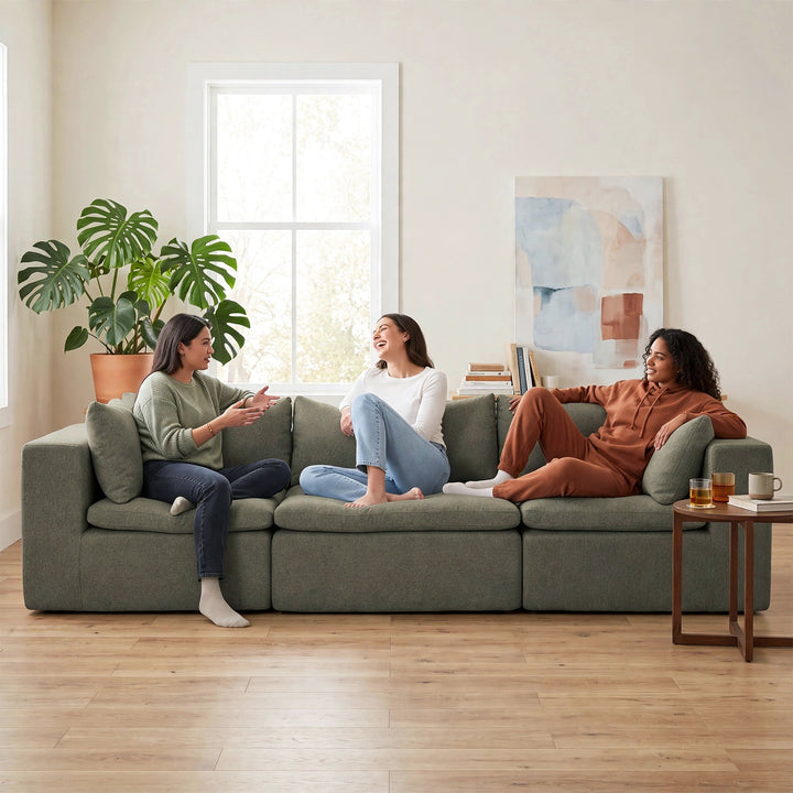 Three women relax on a large sage green sectional sofa in a bright, modern living room with light wood floors, a potted monstera plant, and abstract wall art. 