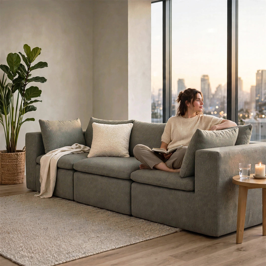 A woman sits on a large gray upholstered sectional sofa in a modern living room with tall windows, a city skyline at dusk, a potted plant, and a small side table with candles. 