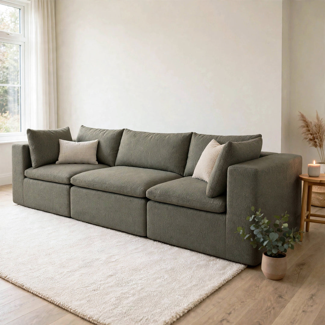 A modern three-seater sofa upholstered in textured gray fabric, styled with two light beige throw pillows in a bright, minimalist living room with a white rug and light wood flooring. 