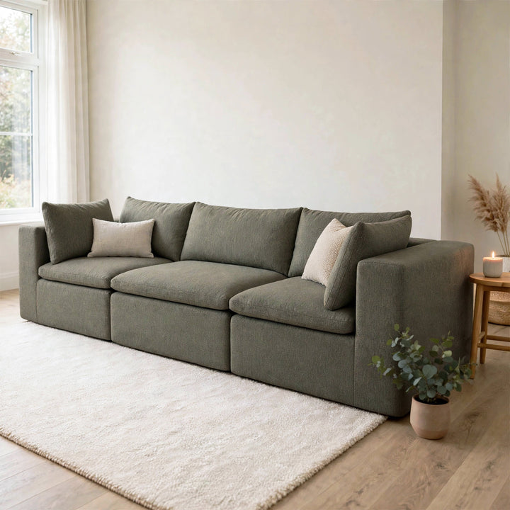 A modern three-seater sofa upholstered in textured gray fabric, styled with two light beige throw pillows in a bright, minimalist living room with a white rug and light wood flooring. 