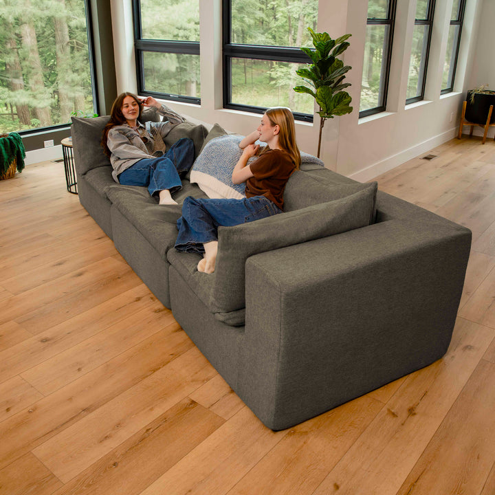 Two women sitting on a large gray sectional sofa in a modern living room with large windows. 