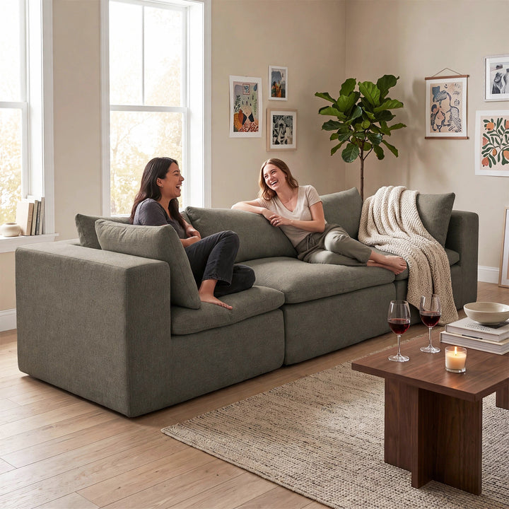A woman and a man relax on a large, modern sectional sofa upholstered in textured gray fabric. The scene is set in a bright, contemporary living room with light wood floors, a wooden coffee table with wine glasses and a candle, framed wall art, and a potted plant. 