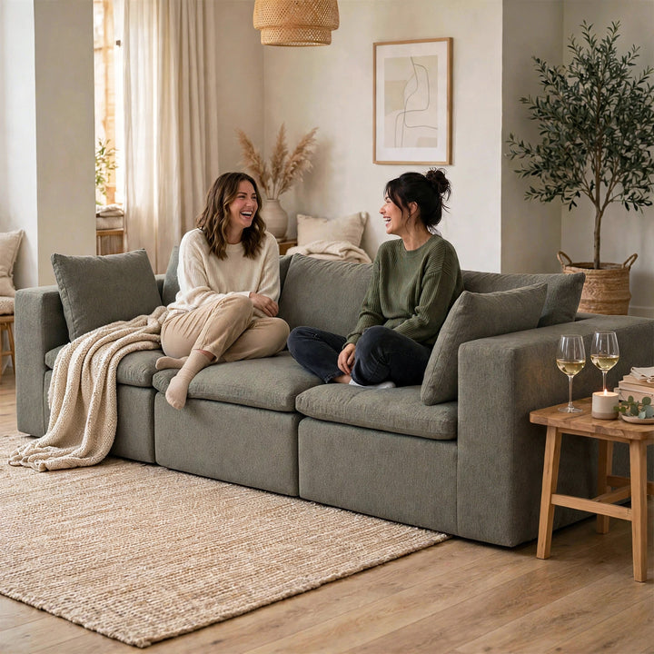 Two women sitting on a large gray fabric sectional sofa in a warm, modern living room. The room features neutral beige and brown tones, with a woven rug, wooden side table, and potted plant. 